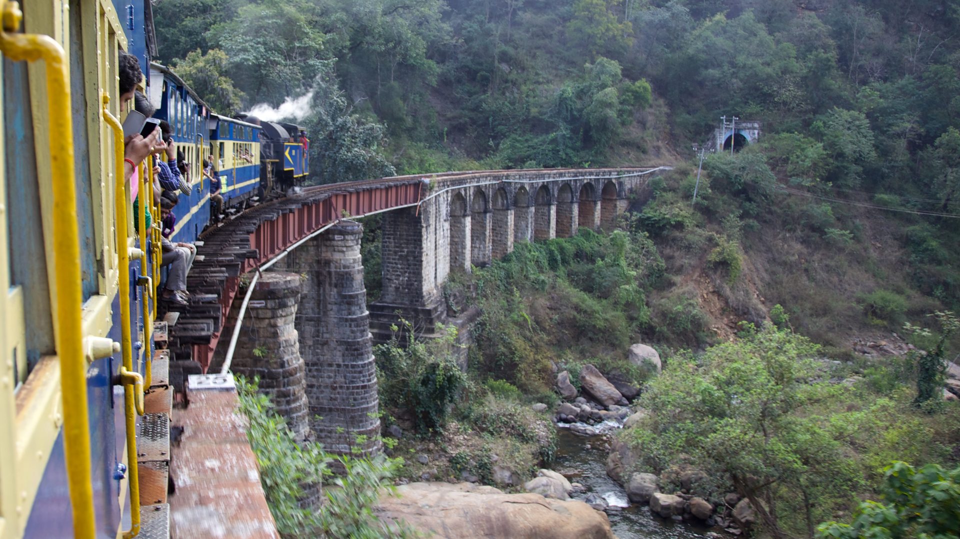 Heritage blue toy train traversing a stone arch viaduct through misty pine-covered Nilgiri hills